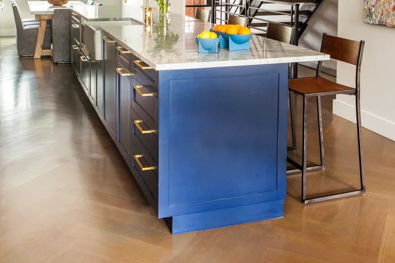 Medium brown wood floor running the length of a kitchen with a blue island and a black steel stair