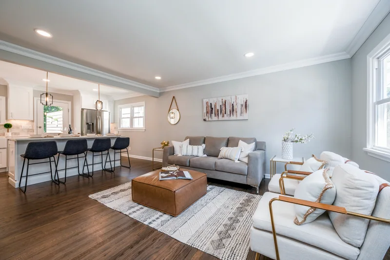 Dark hardwood in an open living room with a tan leather ottoman, kitchen island and stools beyond